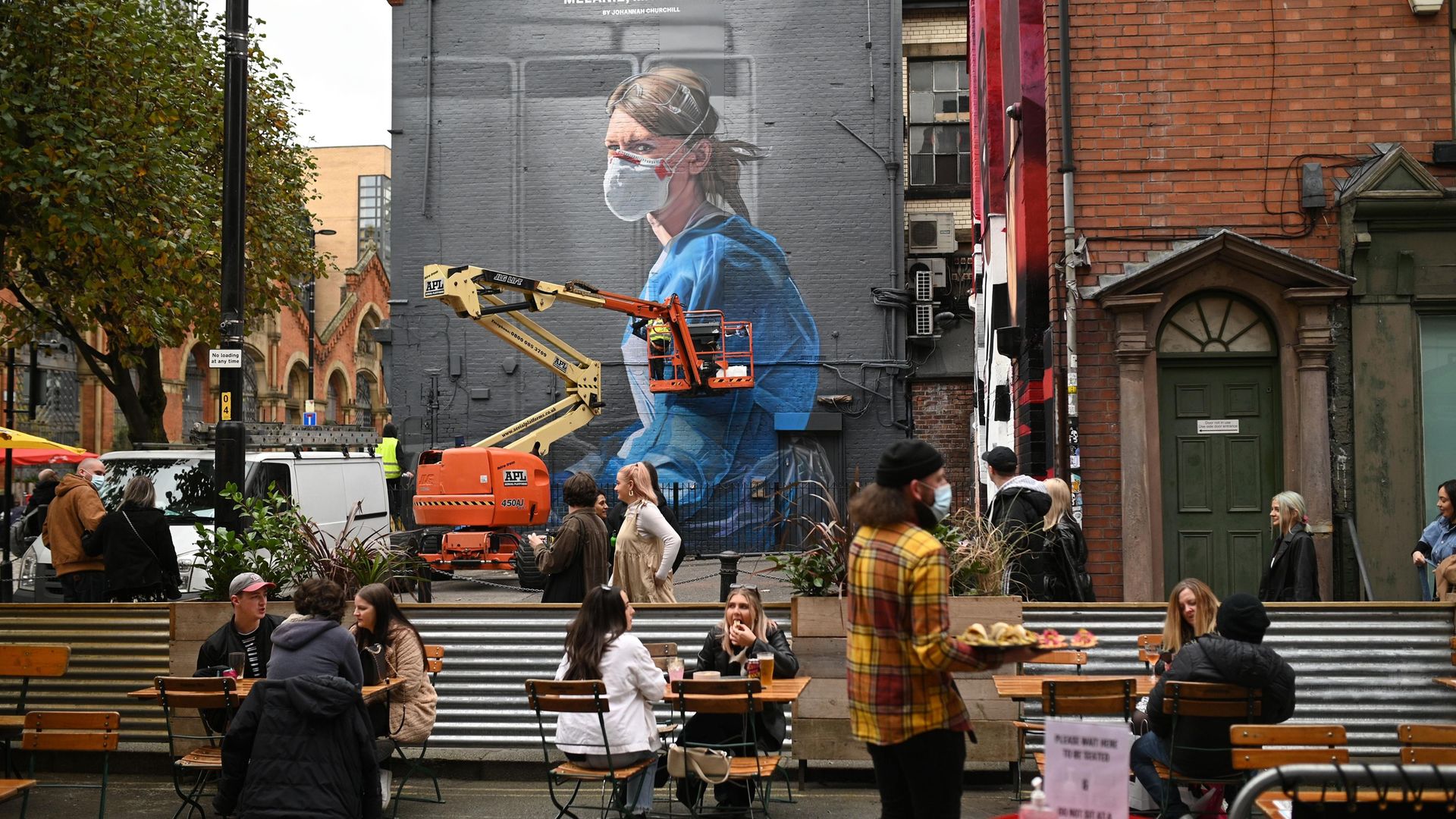People enjoy a socially-distanced drink as an artist creates a mural of a NHS worker on a wall in north Manchester. - Credit: AFP via Getty Images