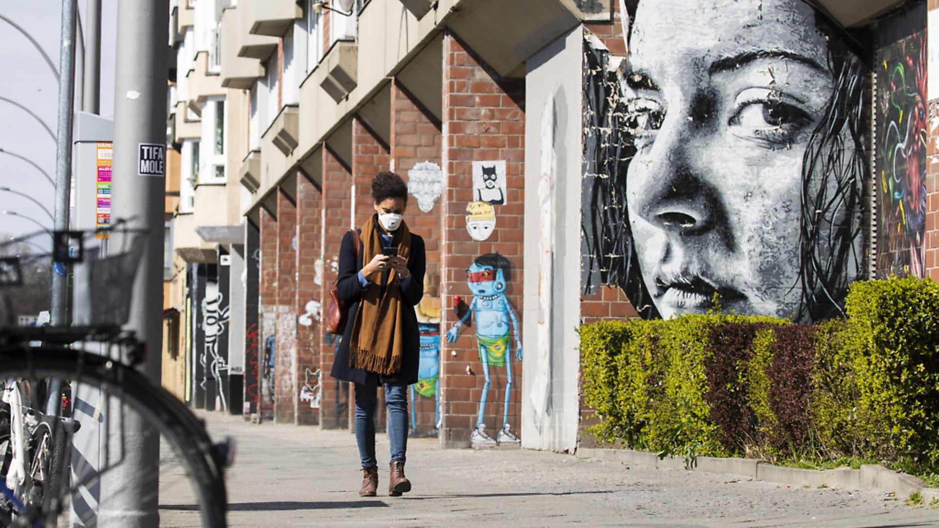 A young woman wearing a protective mask looks at her smartphone while passing by a grafitti representing a watching woman in Berlin, Germany. Picture: Emmanuele Contini/NurPhoto via Getty Images - Credit: NurPhoto via Getty Images