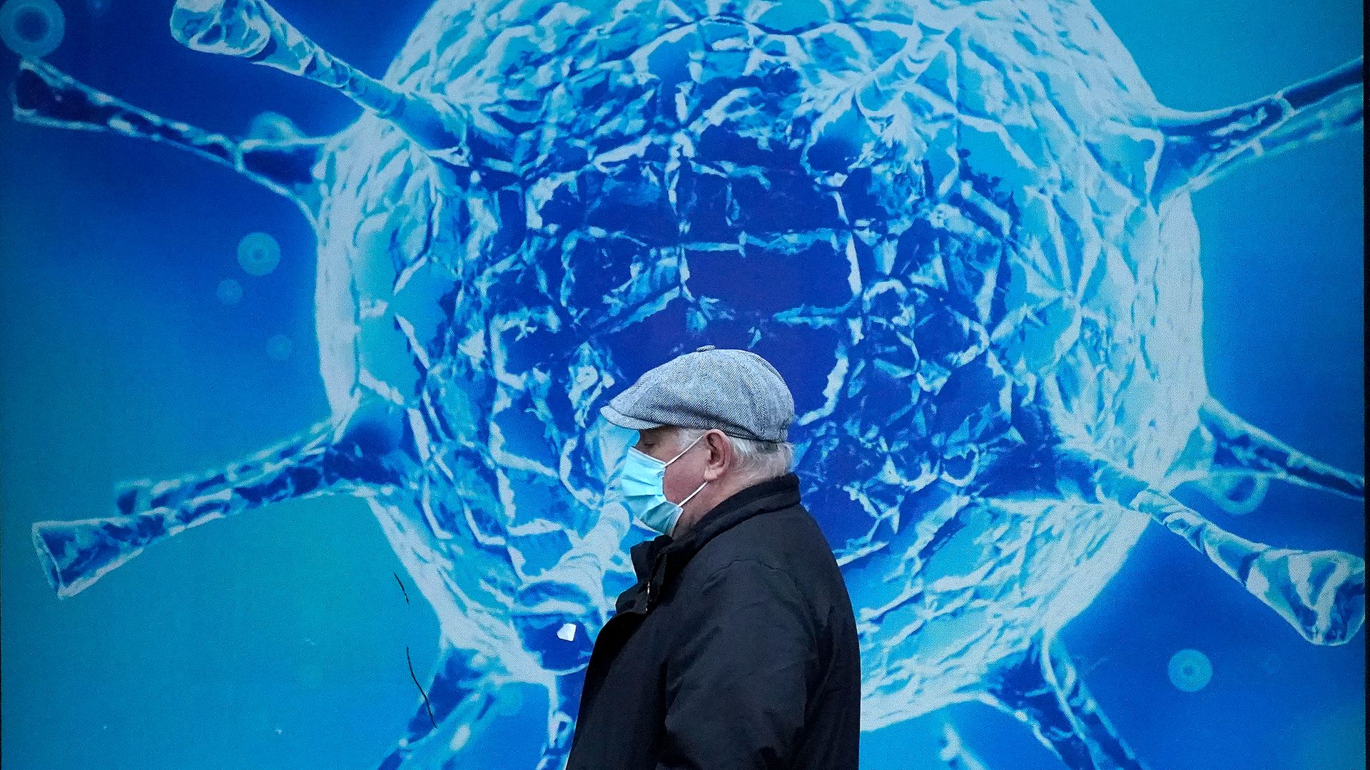 A man wearing a protective face mask walks past an illustration of a virus outside in Oldham - Credit: Getty Images