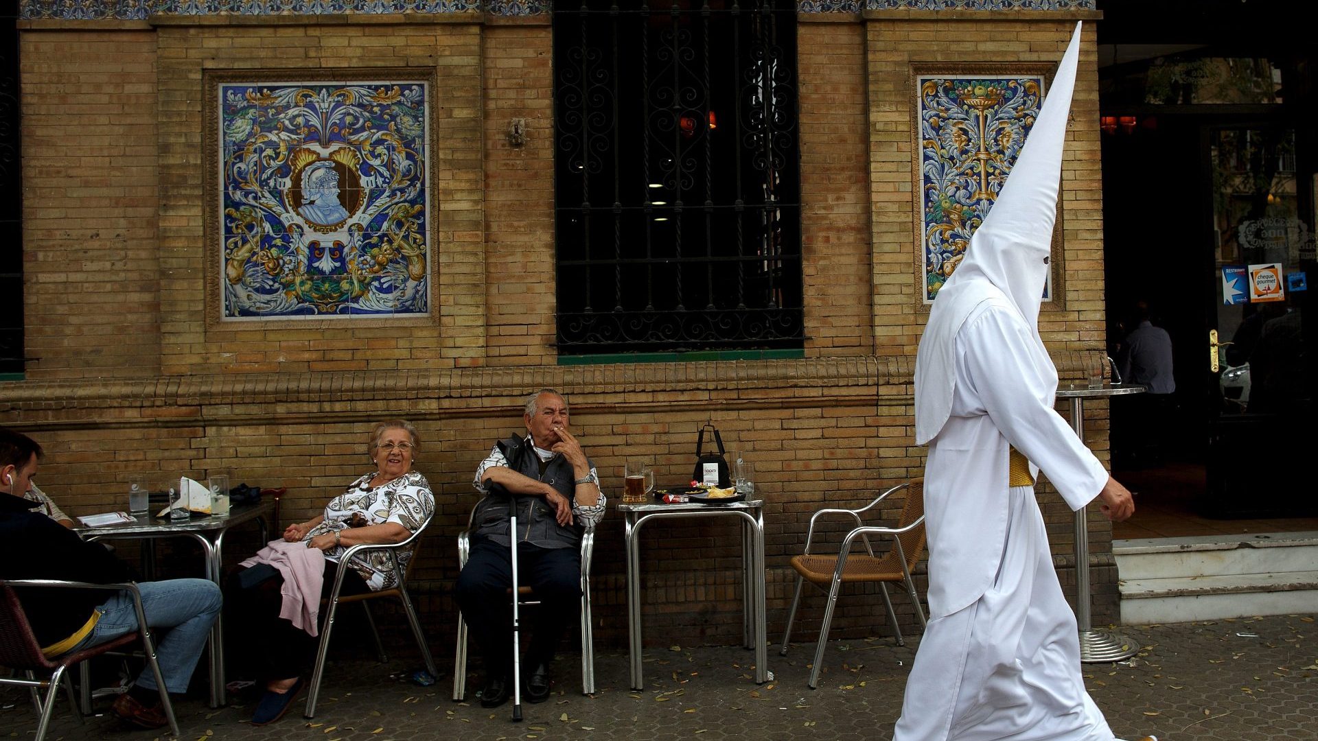 People having tapas outside a bar in Seville are passed by a penitent from the San Gonzalo brotherhood who walks to the church for a procession during Easter week. Photo: Pablo Blázquez Domínguez