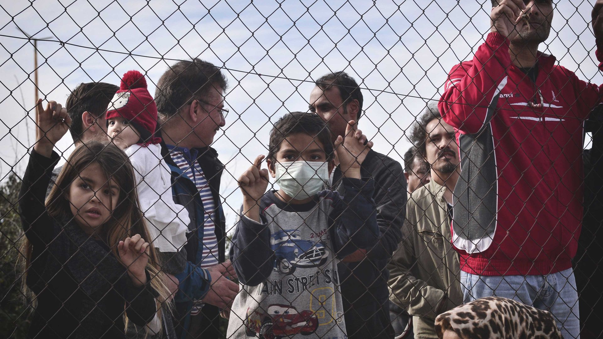 Migrants and children stand behind a fence at the Hellinikon camp in Athens. Photo: LOUISA GOULIAMAKI / AFP